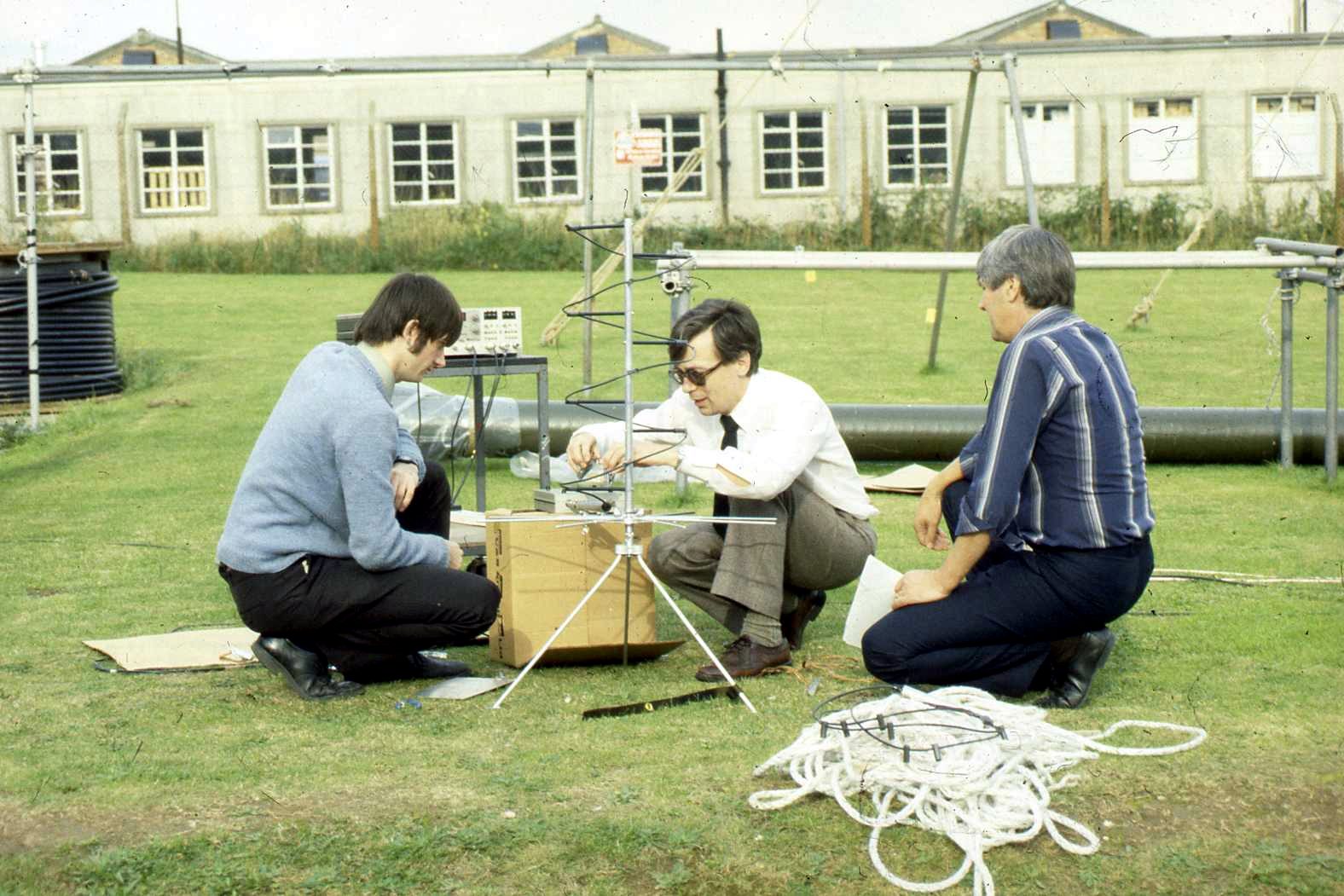 Three engineers kneeling on grass around a helical antenna on a tripod