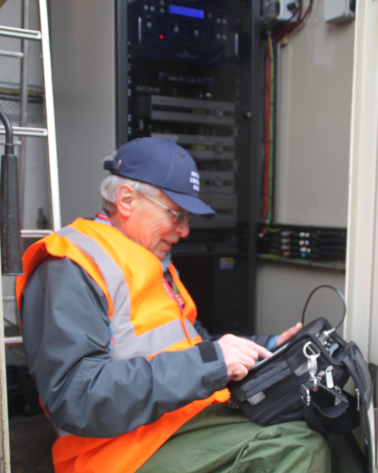 Brian Collins in a hi‑vis vest, seated in an equipment cabinet doorway with a test set